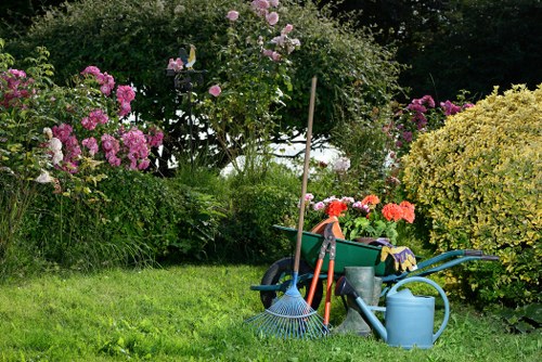 The image depicts a well-maintained garden with a lush green lawn in the foreground, bordered by a variety of flowering shrubs and bushes, including pink and purple blooms, and dense foliage. Behind the plants, there are tall trees providing a natural backdrop. Garden tools such as a blue rake, a watering can, and a bag of compost are placed on the grass, indicating ongoing gardening maintenance. A green wheelbarrow filled with colorful garden supplies, along with a garden fork and a small gardening cloth, are visible near the plants. The garden appears to be part of a residential outdoor space, with a mix of flowering plants and shrubbery that add natural colour and texture, typical of a landscape maintained by professional gardening services in Cricklewood. The scene is set in daylight with clear weather, highlighting the vibrant colours of the flowers and the lushness of the grass, reflecting a healthy and inviting outdoor environment.