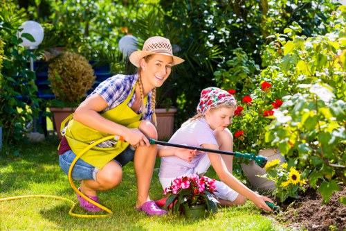 A woman and a young girl are gardening together in a lush outdoor space, likely a front or back garden in Cricklewood. The woman, wearing a straw hat, a yellow apron, and casual clothing, is crouched down with a garden hose in her hand, smiling as she observes the girl. The girl, dressed in a light-colored outfit, a floral hat, and pink gardening shoes, is tending to a flower bed with bright pink, red, and yellow flowers. The garden features a well-maintained lawn with vibrant green grass, bordered by flower beds filled with colorful blooms. Behind them, dense greenery includes trees and shrubs, providing shade and a natural backdrop. Decorative garden lights on tall stakes are visible, adding to the outdoor ambiance. The scene is illuminated by warm sunlight, suggesting a pleasant, sunny day, and the overall environment emphasizes outdoor maintenance and gardening activities typical of a family-managed landscaped yard or front garden in the Cricklewood area. This image aligns with professional gardening services offered by Garden Maintenance Cricklewood, focusing on garden care, planting, and outdoor upkeep.